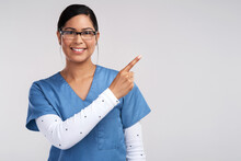 Doctors Eliminate The Nos On Their Vacancy Signs To Exist. Portrait Of A Young Doctor Wearing Glasses And Scrubs, Pointing To Her Left Against A White Background.