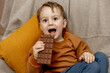 Little adorable boy sitting on the couch at home and eating chocolate bar. Child and sweets, sugar confectionery. Kid enjoy a delicious dessert. Preschool child with casual clothing. Positive emotion.