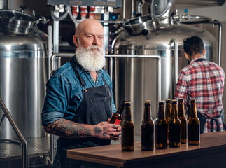 Photo of two elderly brewers working in beer factory with barrels and table there are beer bottles.