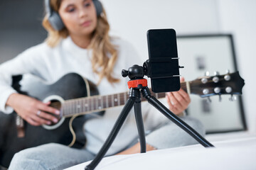 Poster - My followers requested this song. Shot of a young woman using her cellphone to record herself while playing the guitar at home.