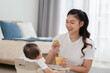 © 220 Selfmade studio - Mother feeding her asian baby daughter with pumpkin mashed or vegetable mash for vitamin on rubber spoon.Mom trying feed little baby boy at home enjoy and spending time together.Baby feeding Concept