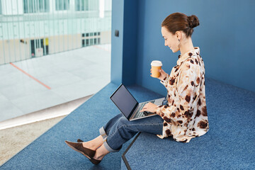 Graphic portrait of young professional using laptop and enjoying coffee while working in blue office space