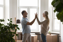 Mature Couple Accomplish Relocation Day Give High Five Feel Happy, Standing In Unfurnished Living Room With Heap Of Cardboard Boxes. Move Celebration, Bank Mortgage For Older Citizen, New Life Concept