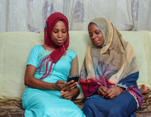 Wall Mural - Two happy African Nigerian female muslim sisters or friends with Hijab scarf, sitting together as they make use of a smart phone with them