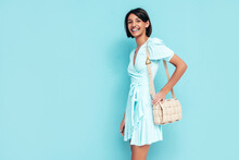 Portrait Of Young Beautiful Smiling Female In Trendy Summer Dress. Carefree Woman Posing Near Blue Wall In Studio. Positive Model Having Fun Indoors With Handbag. Cheerful And Happy. Isolated