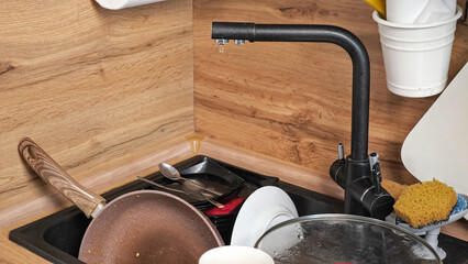 Heap of dirty dishes after cooking dinner lies in kitchen. Broken faucet leaks dropping water on utensils in sink at wooden countertop close-up