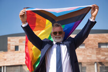 Mature Gay Man, Executive, Gray-haired, With Beard, Sunglasses, Jacket And Tie, Waving The New Lgbtiq  Pride Flag In The Wind Under A Blue Sky. Concept Mature Gay Man, Lgbt, Pride, Teddy Bear.