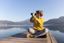 A Boy With Binoculars Sitting On A Wooden Pier And Looking On The Mountains. Hiking Outdoor Concept