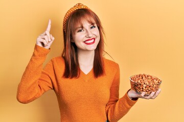 Canvas Print - Redhead young woman holding peanuts smiling with an idea or question pointing finger with happy face, number one