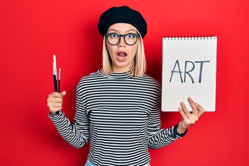 Poster - Beautiful blonde woman holding art notebook and brushes afraid and shocked with surprise and amazed expression, fear and excited face.