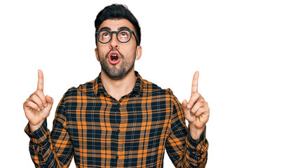 Wall Mural - Hispanic man with beard wearing casual clothes and glasses amazed and surprised looking up and pointing with fingers and raised arms.