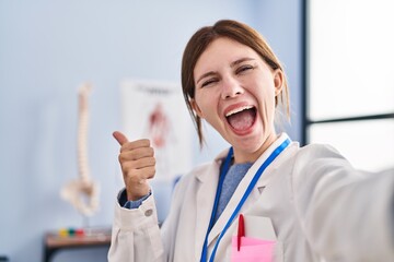 Sticker - Young physiotherapist woman working at pain recovery clinic pointing thumb up to the side smiling happy with open mouth