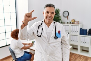 Poster - Middle age doctor man at the clinic with a patient smiling and confident gesturing with hand doing small size sign with fingers looking and the camera. measure concept.