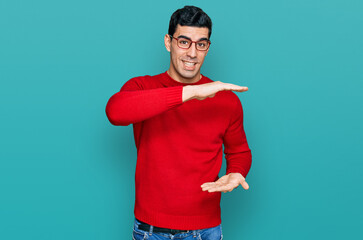 Wall Mural - Handsome hispanic man wearing casual clothes and glasses gesturing with hands showing big and large size sign, measure symbol. smiling looking at the camera. measuring concept.