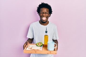 Poster - Young african american man holding tray with breakfast food sticking tongue out happy with funny expression.