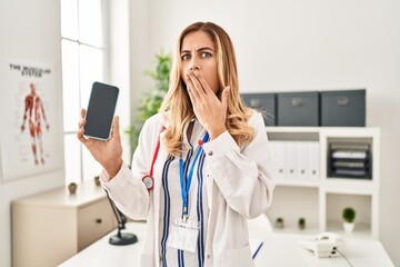 Canvas Print - Young blonde doctor woman working at the clinic showing smartphone screen covering mouth with hand, shocked and afraid for mistake. surprised expression