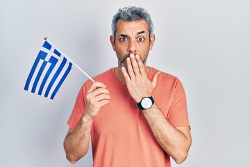 Canvas Print - Handsome middle age man with grey hair holding greece flag covering mouth with hand, shocked and afraid for mistake. surprised expression