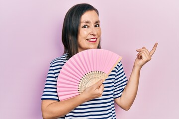 Poster - Middle age hispanic woman waving hand fan cooling air in summer smiling happy pointing with hand and finger to the side