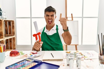 Poster - Young hispanic man at art studio holding degree angry and mad raising fist frustrated and furious while shouting with anger. rage and aggressive concept.