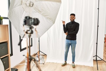 Wall Mural - Young hispanic man with beard posing as model at photography studio with a big smile on face, pointing with hand and finger to the side looking at the camera.