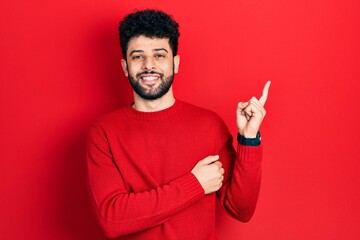 Wall Mural - Young arab man with beard wearing casual red sweater with a big smile on face, pointing with hand and finger to the side looking at the camera.