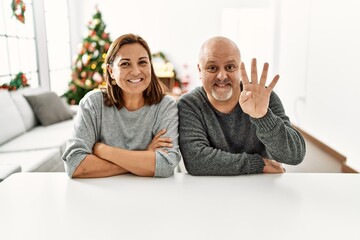 Wall Mural - Middle age hispanic couple sitting on the table by christmas tree showing and pointing up with fingers number four while smiling confident and happy.