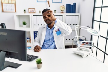 Wall Mural - Young african doctor man wearing stethoscope at the clinic smiling happy doing ok sign with hand on eye looking through fingers