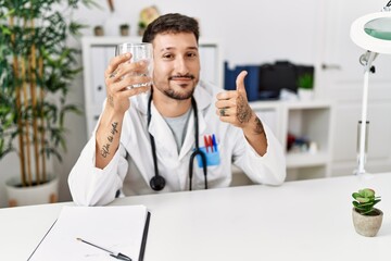 Wall Mural - Young doctor holding glass of water smiling happy and positive, thumb up doing excellent and approval sign