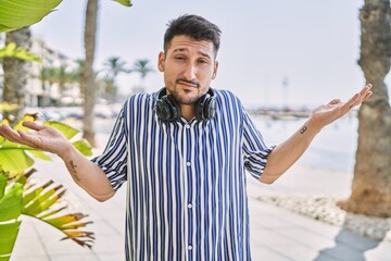 Wall Mural - Young handsome man listening to music using headphones outdoors clueless and confused expression with arms and hands raised. doubt concept.