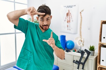 Poster - Young man with beard working at pain recovery clinic smiling making frame with hands and fingers with happy face. creativity and photography concept.