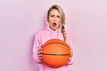 Canvas Print - Beautiful young blonde woman holding basketball ball in shock face, looking skeptical and sarcastic, surprised with open mouth