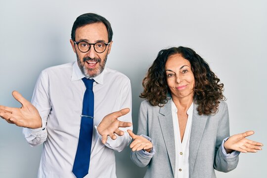 Middle age couple of hispanic woman and man wearing business office uniform clueless and confused expression with arms and hands raised. doubt concept.