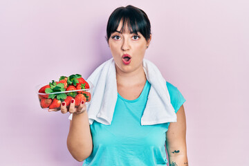 Canvas Print - Young hispanic woman wearing sportswear holding strawberries scared and amazed with open mouth for surprise, disbelief face