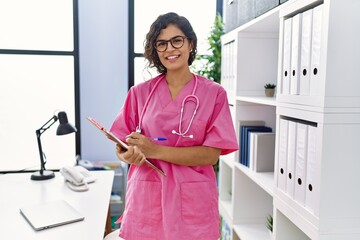 Wall Mural - Young latin woman wearing doctor uniform writing on clipboard at clinic