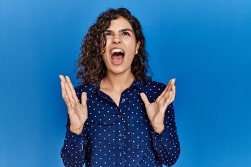 Poster - Young brunette woman with curly hair wearing casual clothes over blue background crazy and mad shouting and yelling with aggressive expression and arms raised. frustration concept.