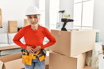 Canvas Print - Middle age grey-haired woman wearing hardhat standing at new home with hand on stomach because indigestion, painful illness feeling unwell. ache concept.