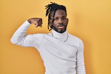 Poster - African man with dreadlocks wearing turtleneck sweater over yellow background strong person showing arm muscle, confident and proud of power