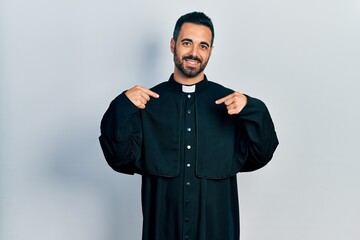 Sticker - Handsome hispanic man with beard wearing catholic priest robe looking confident with smile on face, pointing oneself with fingers proud and happy.