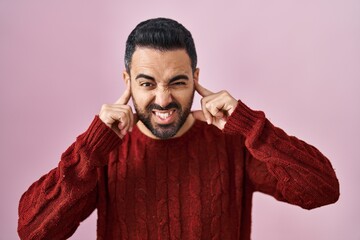 Poster - Young hispanic man with beard wearing casual sweater over pink background covering ears with fingers with annoyed expression for the noise of loud music. deaf concept.