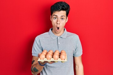 Young hispanic man showing fresh white eggs scared and amazed with open mouth for surprise, disbelief face