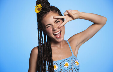 Portrait of happy smiling young woman with tan, summer flower in hair, showing peace, v-sign, concept of vacation and holidays, standing over blue background