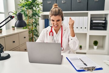 Canvas Print - Young doctor woman wearing doctor uniform working using computer laptop annoyed and frustrated shouting with anger, yelling crazy with anger and hand raised