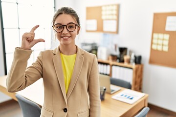 Canvas Print - Young brunette teenager wearing business style at office smiling and confident gesturing with hand doing small size sign with fingers looking and the camera. measure concept.