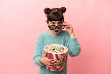 Wall Mural - Little caucasian girl isolated on pink background surprised with 3d glasses and holding a big bucket of popcorns