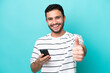 © luismolinero - Young Brazilian man isolated on blue background using mobile phone while doing thumbs up