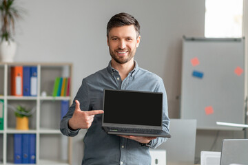 Sticker - Portrait of happy mature businessman pointing at laptop computer with blank black screen, standing in office, mockup