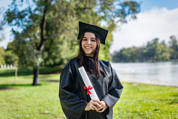 Happy caucasian graduated girl with long brown hair. She is wearing a bachelor gown and a black mortarboard. She is holding a diploma