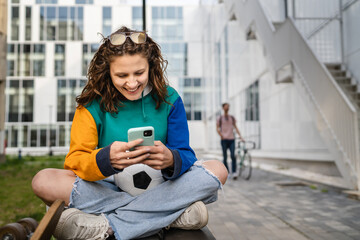 One young caucasian woman female sitting on the bench in front of the building or at stadium with soccer ball waiting for the football game using mobile phone real people copy space