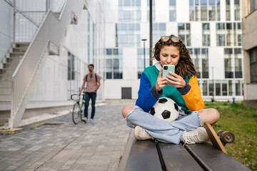 One young caucasian woman female sitting on the bench in front of the building or at stadium with soccer ball waiting for the football game using mobile phone real people copy space