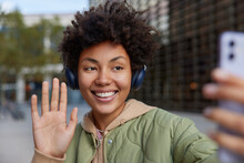 Cheerful Curly Woman Takes Self Portrait Via Smartphone Waves Palm Says Hello Listens Music Via Headphones Smiles Gladfully Poses Outdoors Against Blurred Background Has Recreation Using Technology